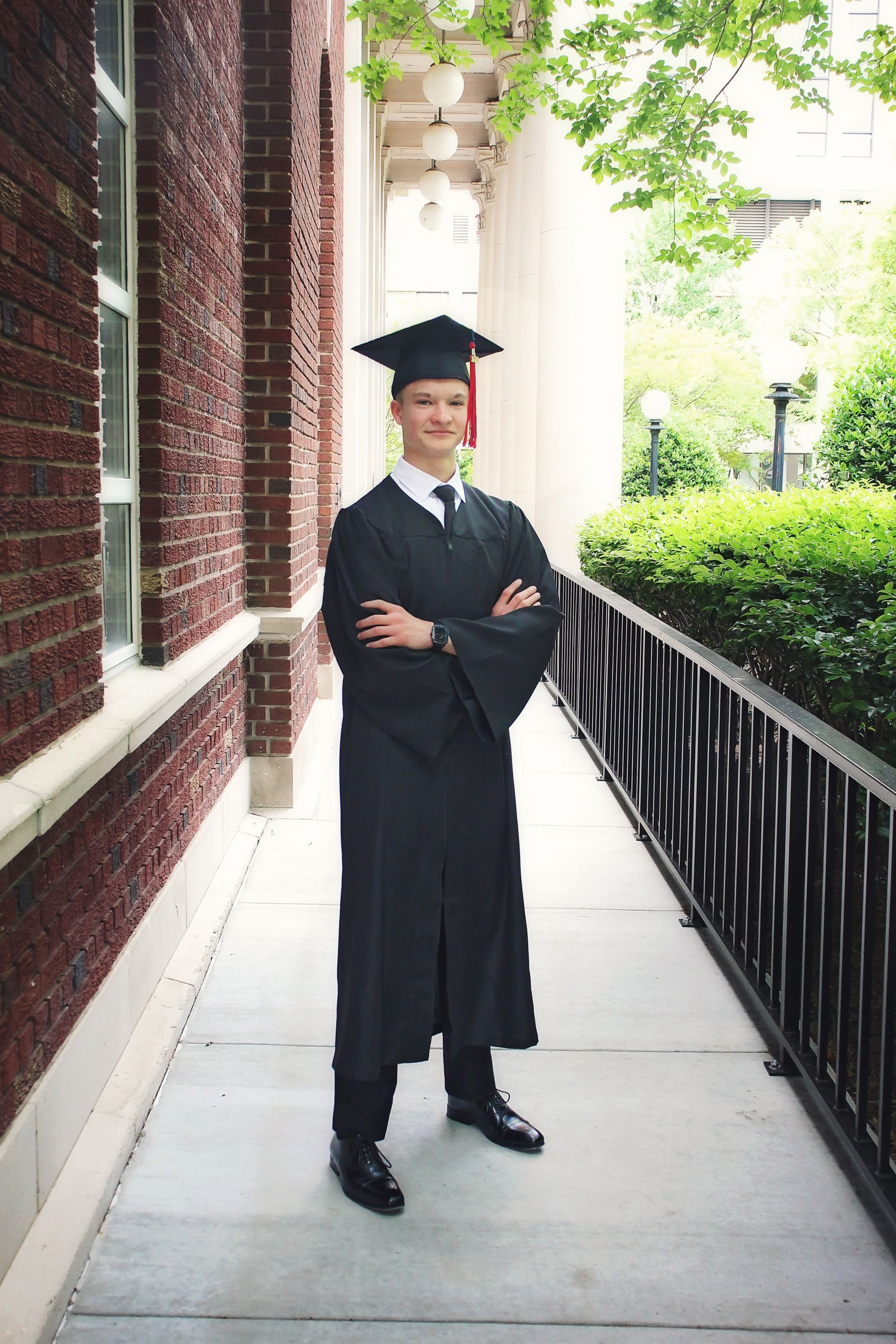 A man in a graduation cap and gown is standing on a sidewalk with his arms crossed.