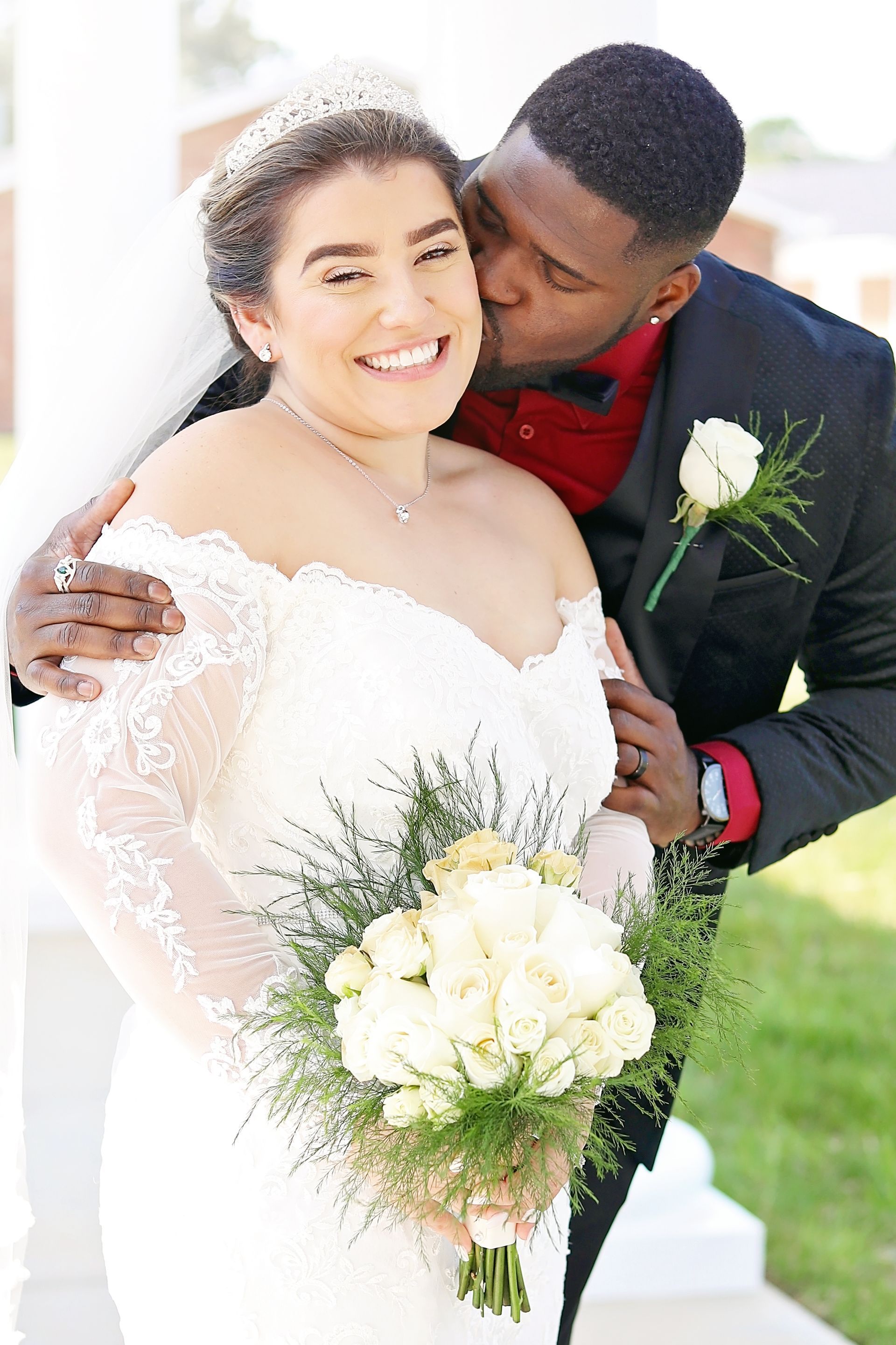 A bride and groom are posing for a picture and the groom is kissing the bride on the cheek.