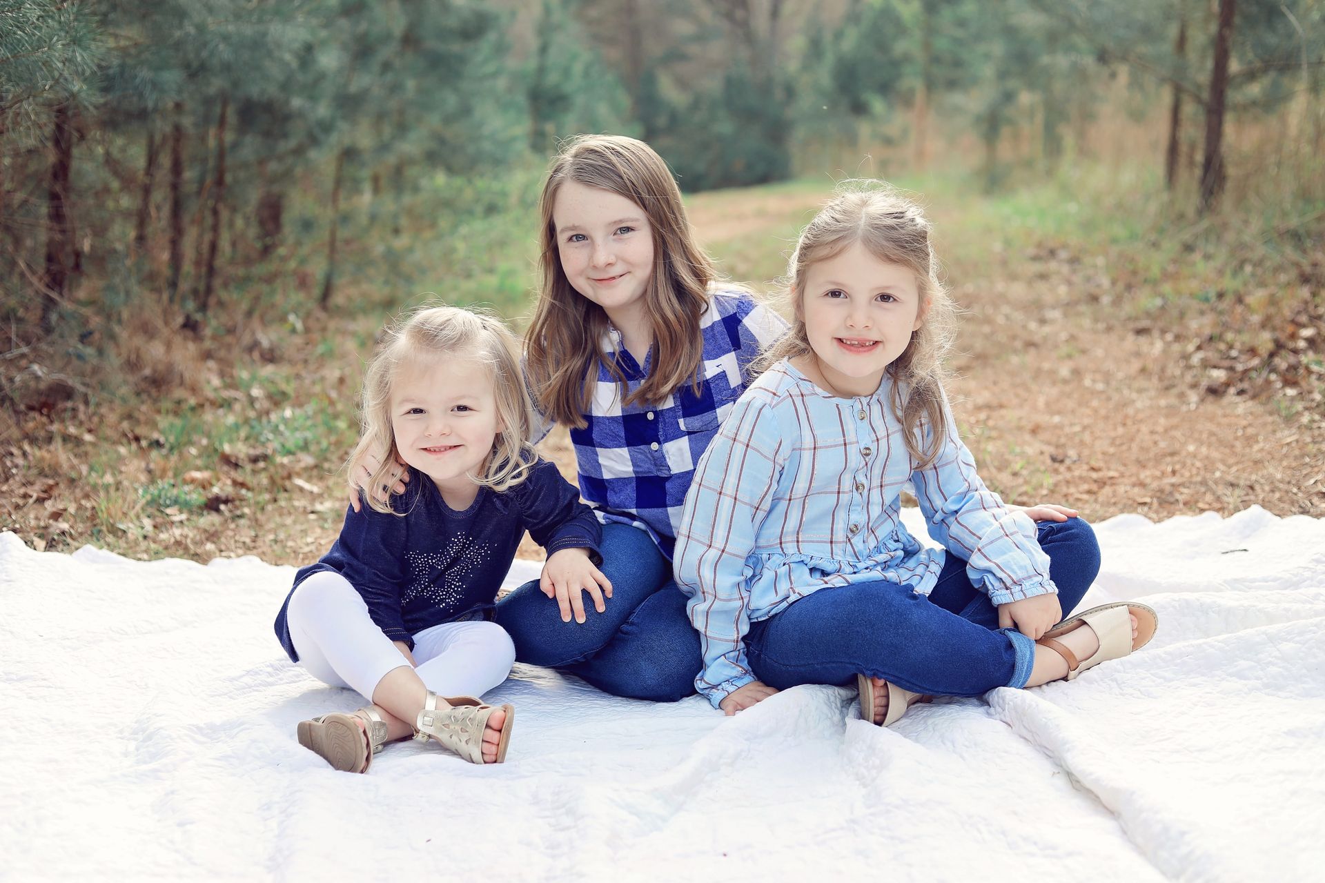 Three little girls are sitting on a blanket in the woods.