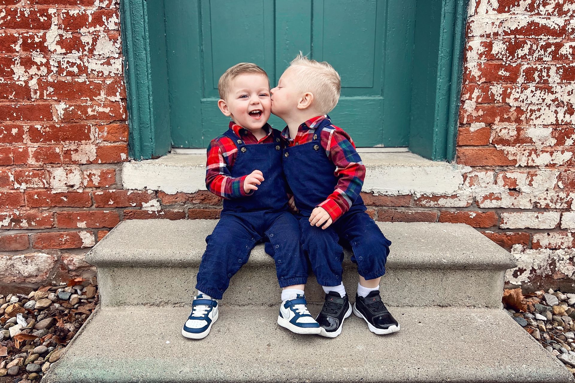 Two young boys are kissing each other on the cheek while sitting on steps in front of a brick building.