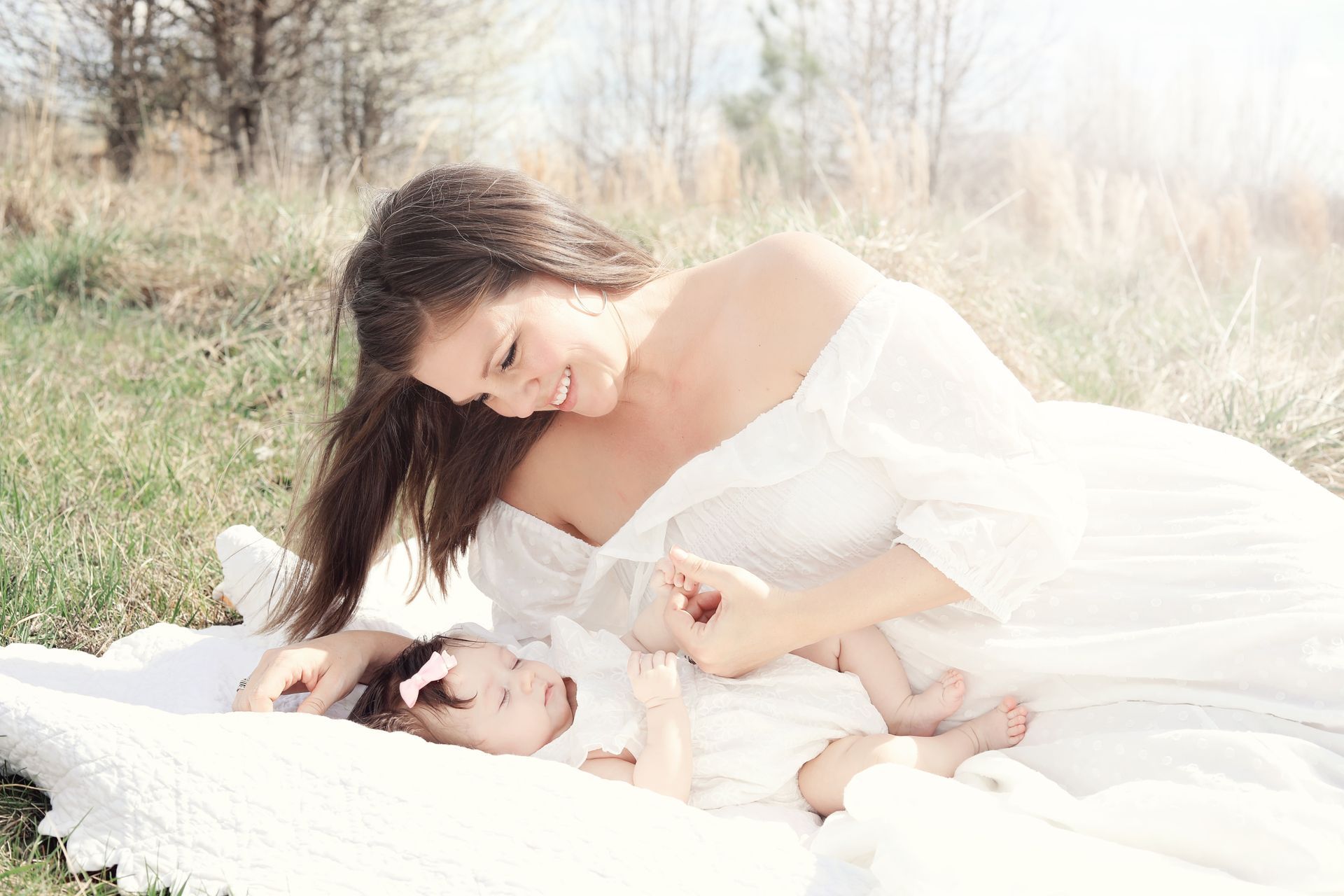 A woman in a white dress is laying on a blanket with a baby.