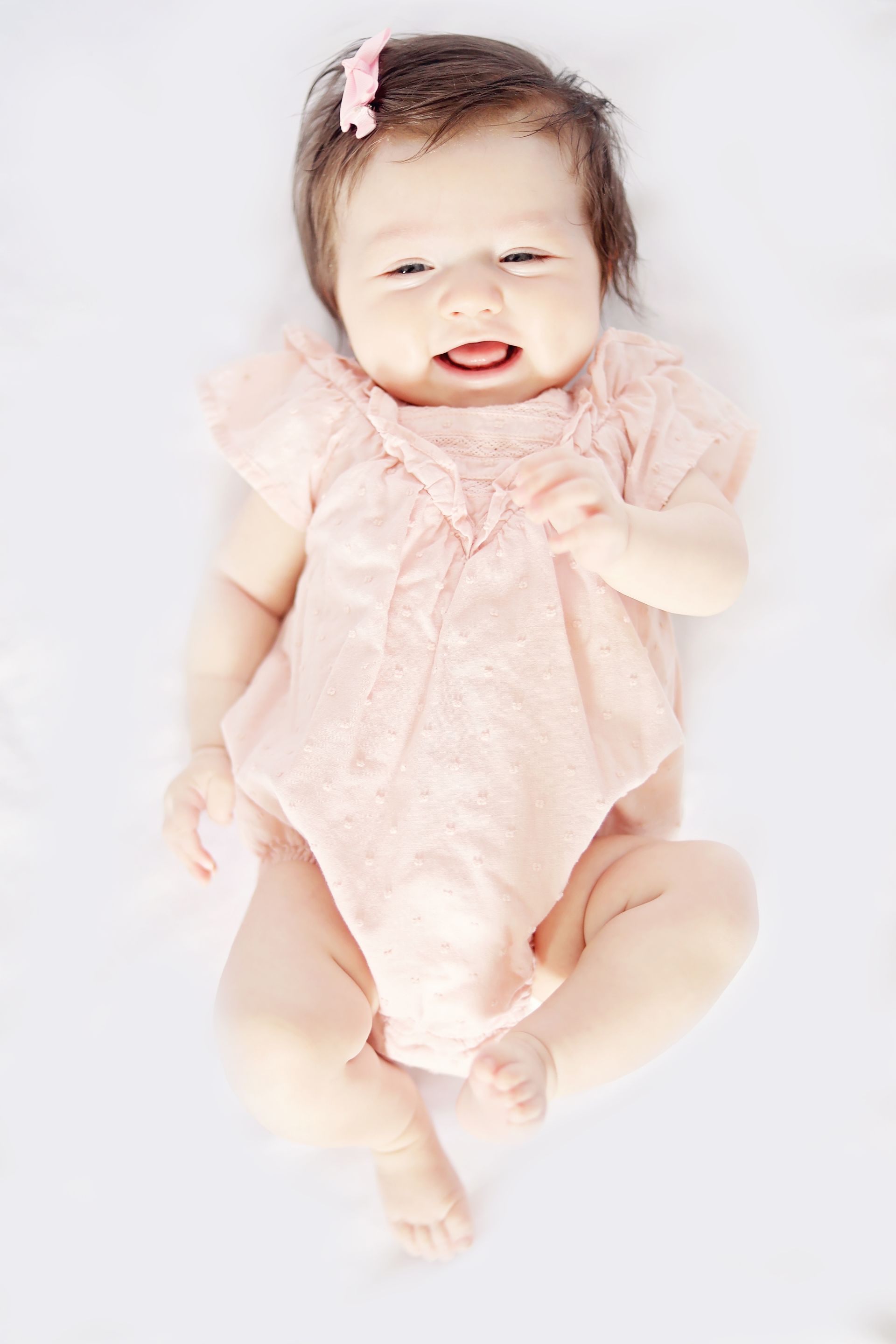 A baby girl in a pink dress is laying on a white blanket and smiling.