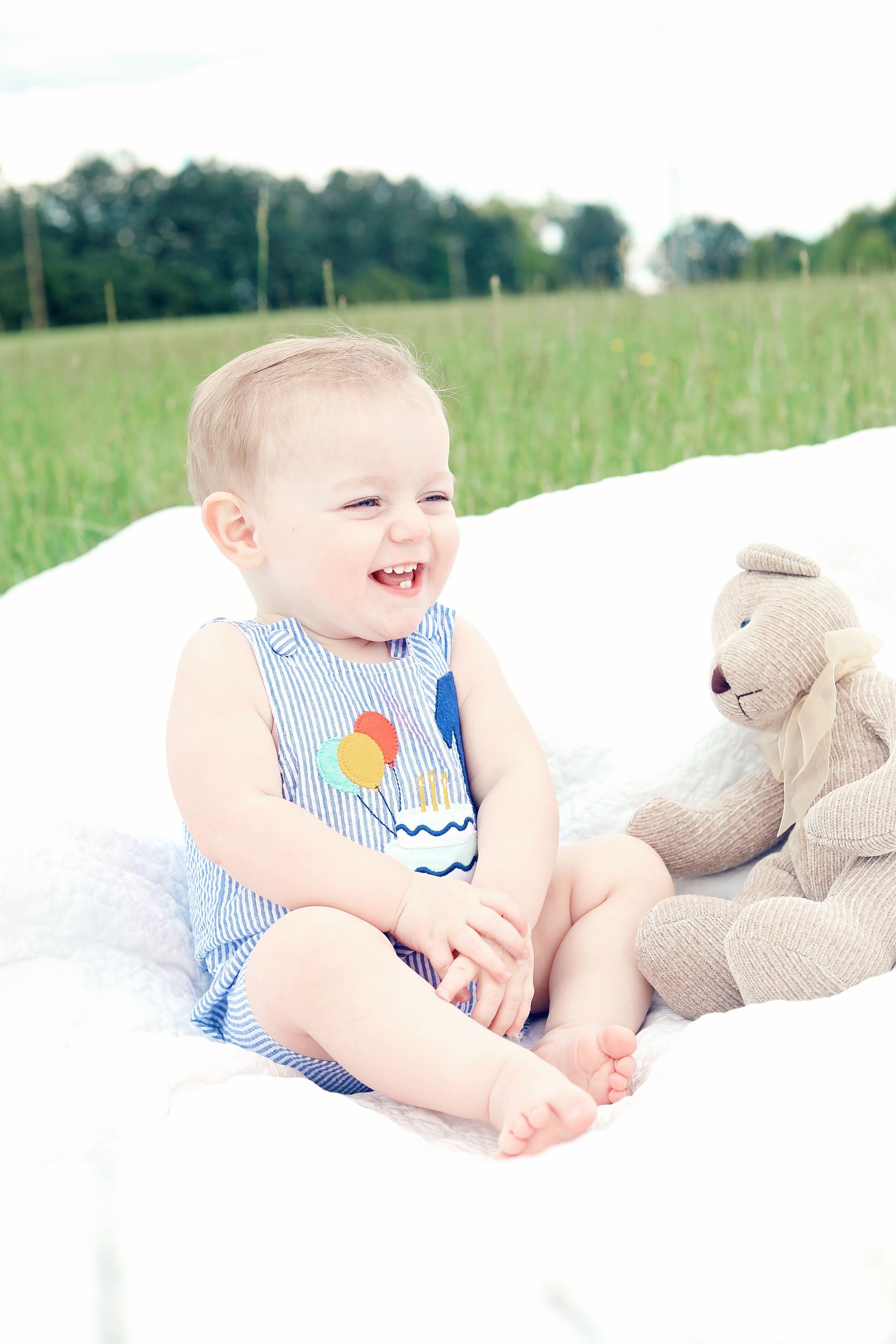 A baby is sitting on a blanket next to a teddy bear.