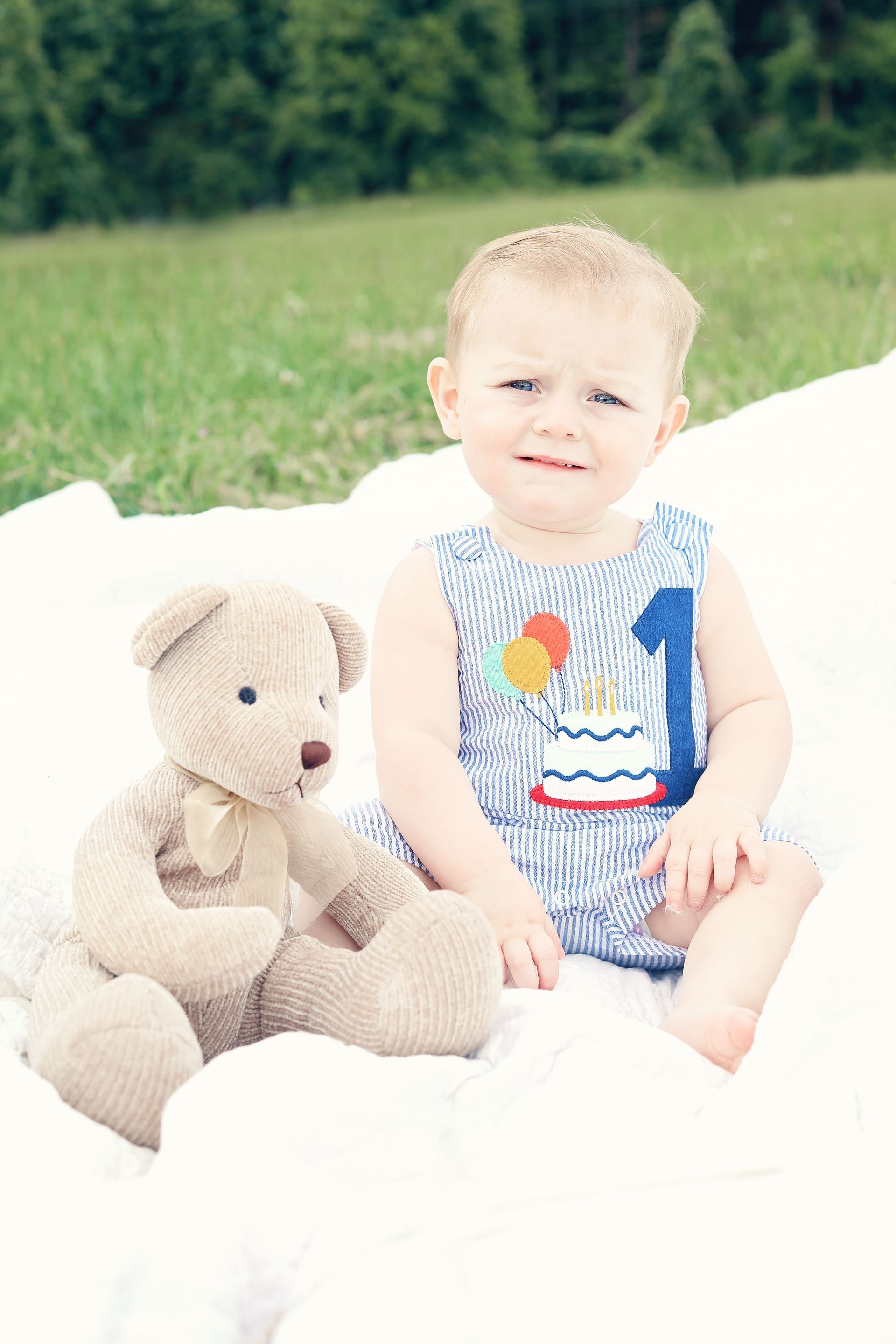 A baby is sitting next to a teddy bear on a white blanket.