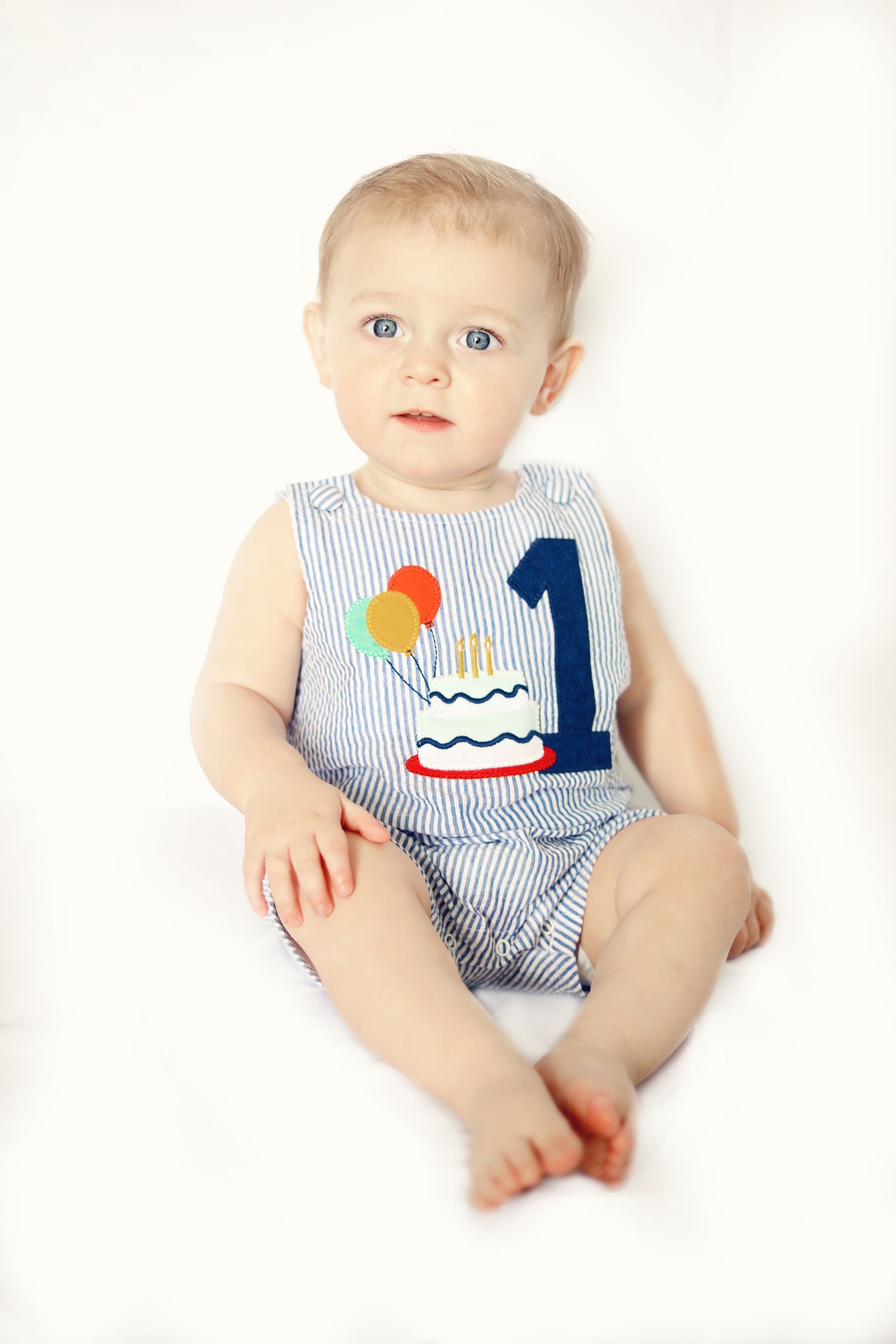 A baby is wearing a birthday outfit with a cake and balloons on it.