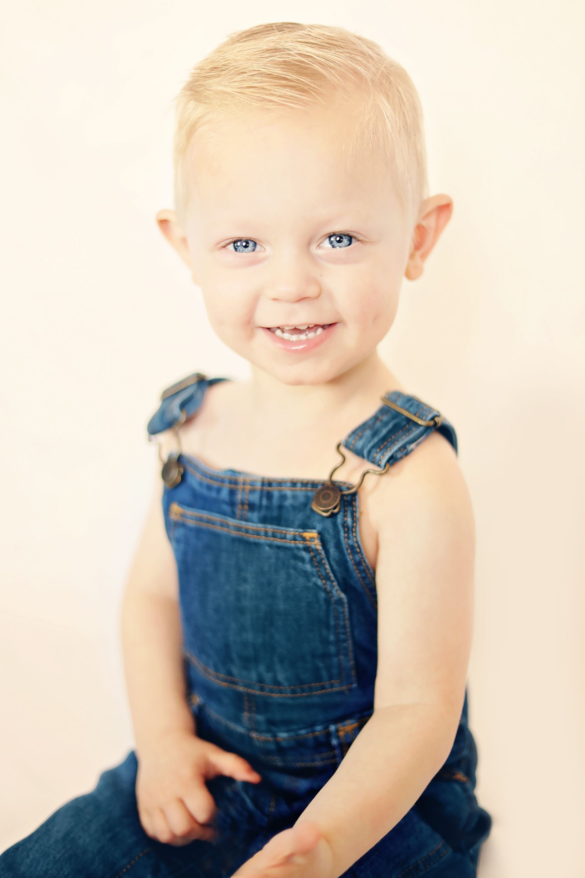 A young boy wearing overalls is sitting on a white surface.