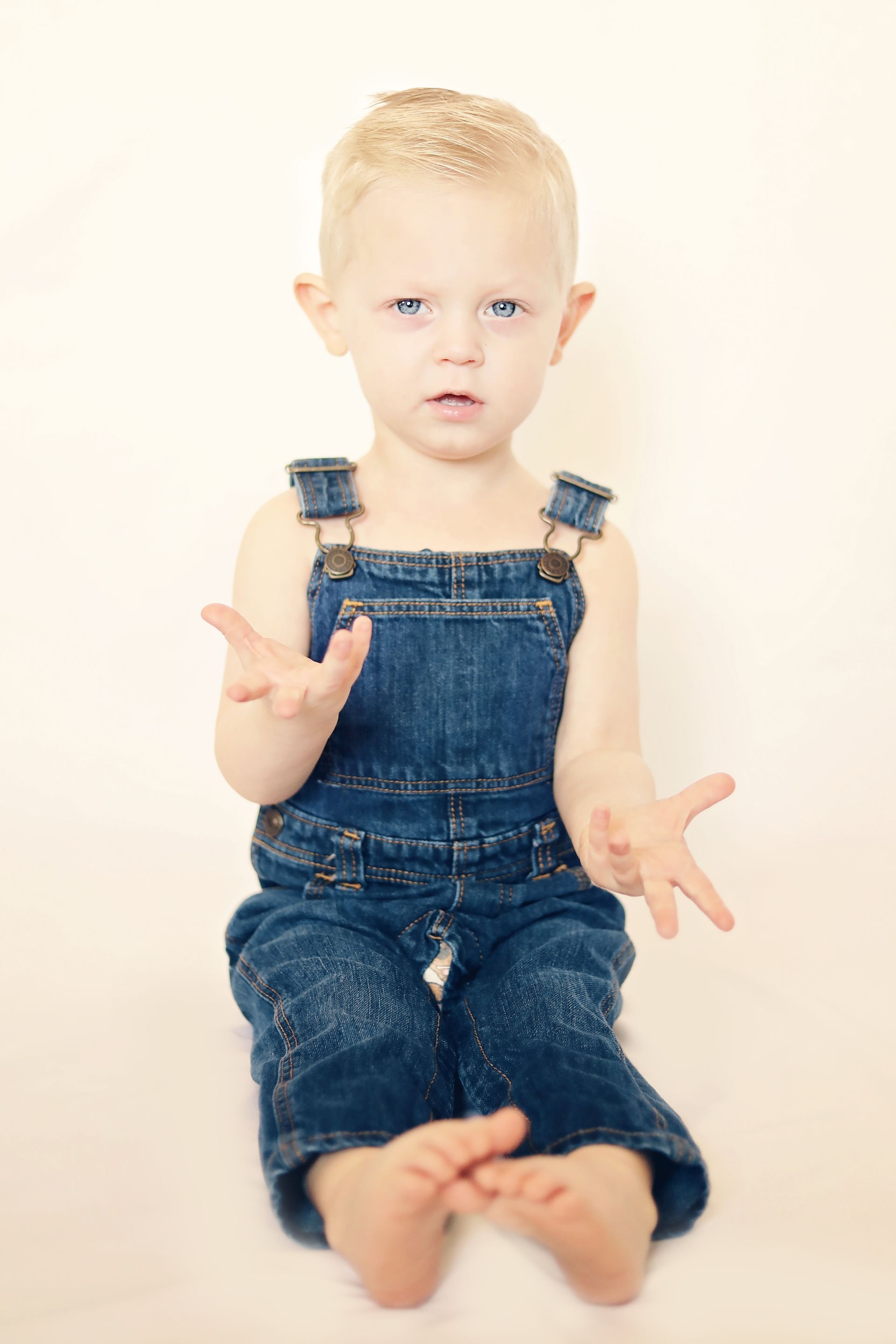 A young boy wearing overalls is sitting on the floor.