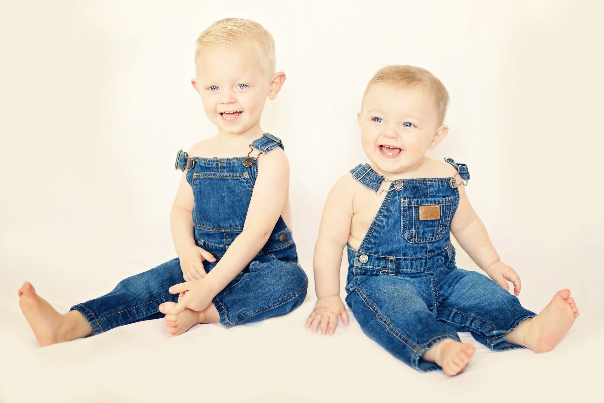 Two babies wearing overalls are sitting next to each other on a white surface.