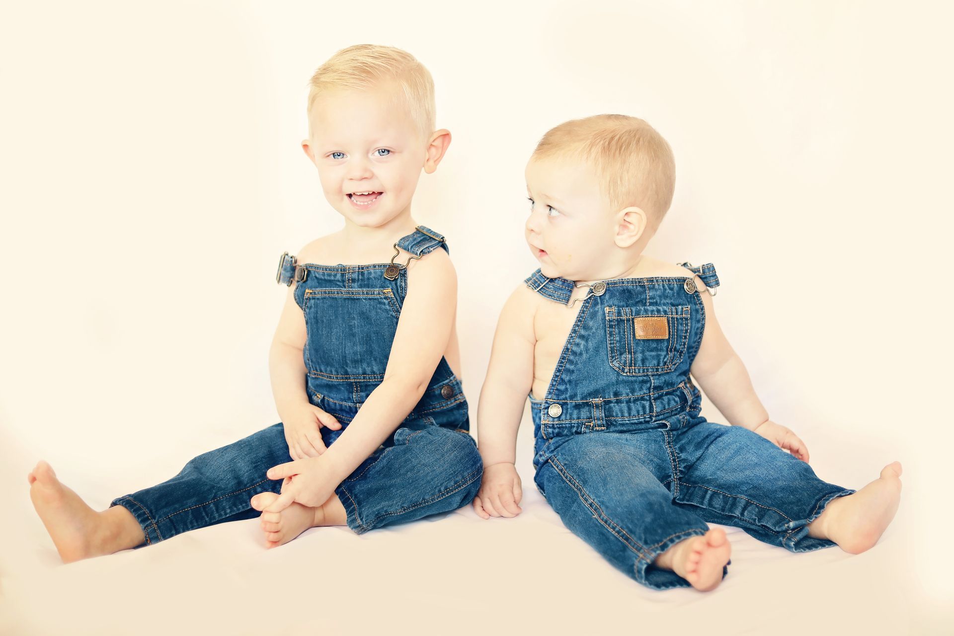 Two young boys in overalls are sitting next to each other on the floor.