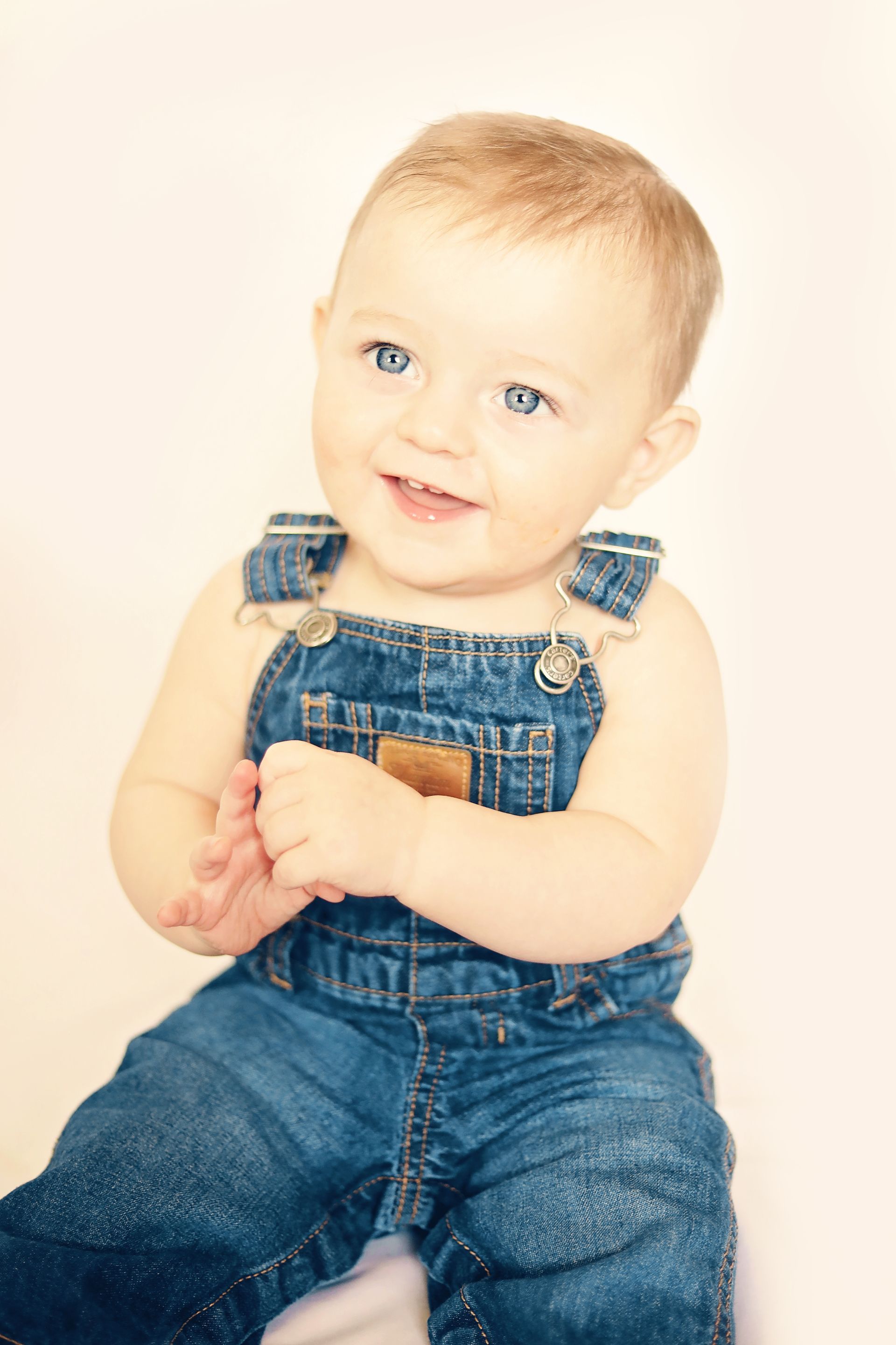A baby in overalls is sitting on a white surface and smiling.