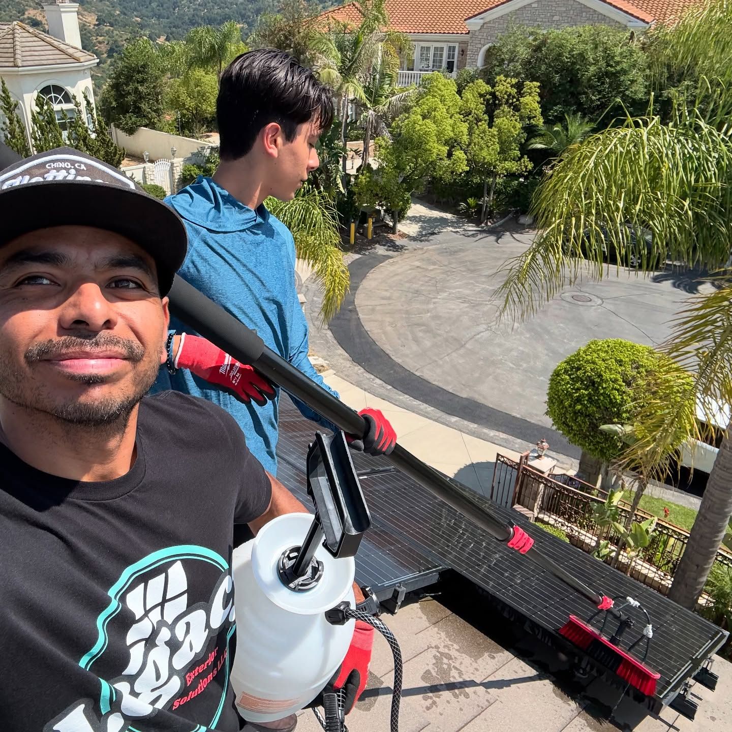 Man and teen on a roof cleaning a solar panel with a sprayer and long-handled brush, sunny day.