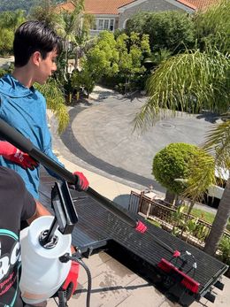 Person using a long-handled cleaning tool on a rooftop, with red gloves. In the background, a driveway and trees are visible.
