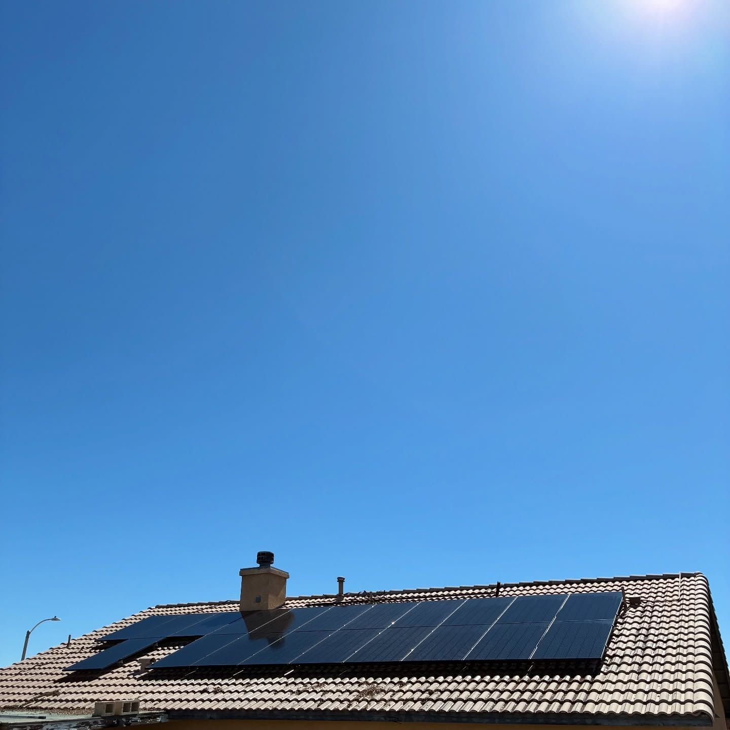 Solar panels on a residential roof under a bright blue sky, with the sun in the upper right corner.