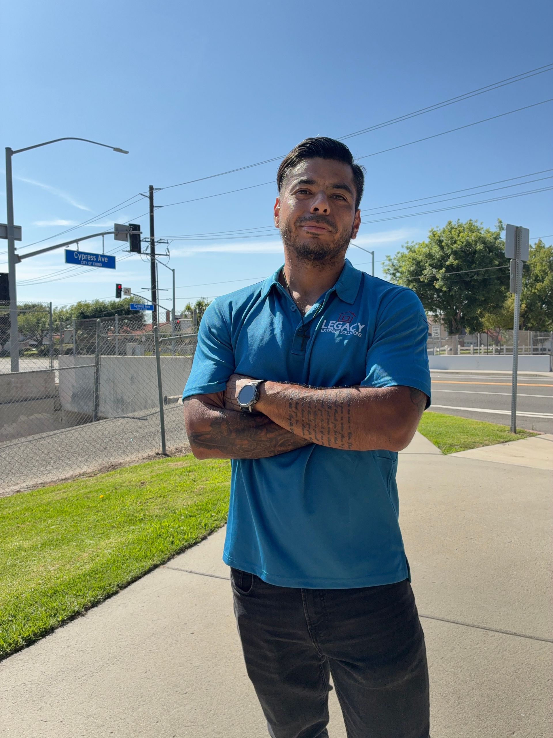 Man with crossed arms standing on a sidewalk in front of a crosswalk, wearing a blue shirt and dark pants under a bright blue sky.
