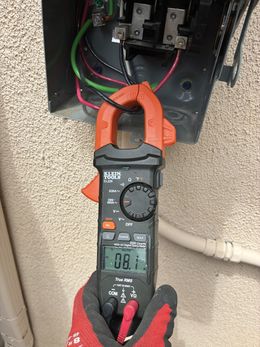 A person uses a clamp meter to measure electrical current inside a metal box on a stucco wall. The meter reads 0.1 amps.