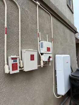 Exterior wall with electrical boxes, conduits, and a solar panel inverter. White boxes and conduits against gray stucco.