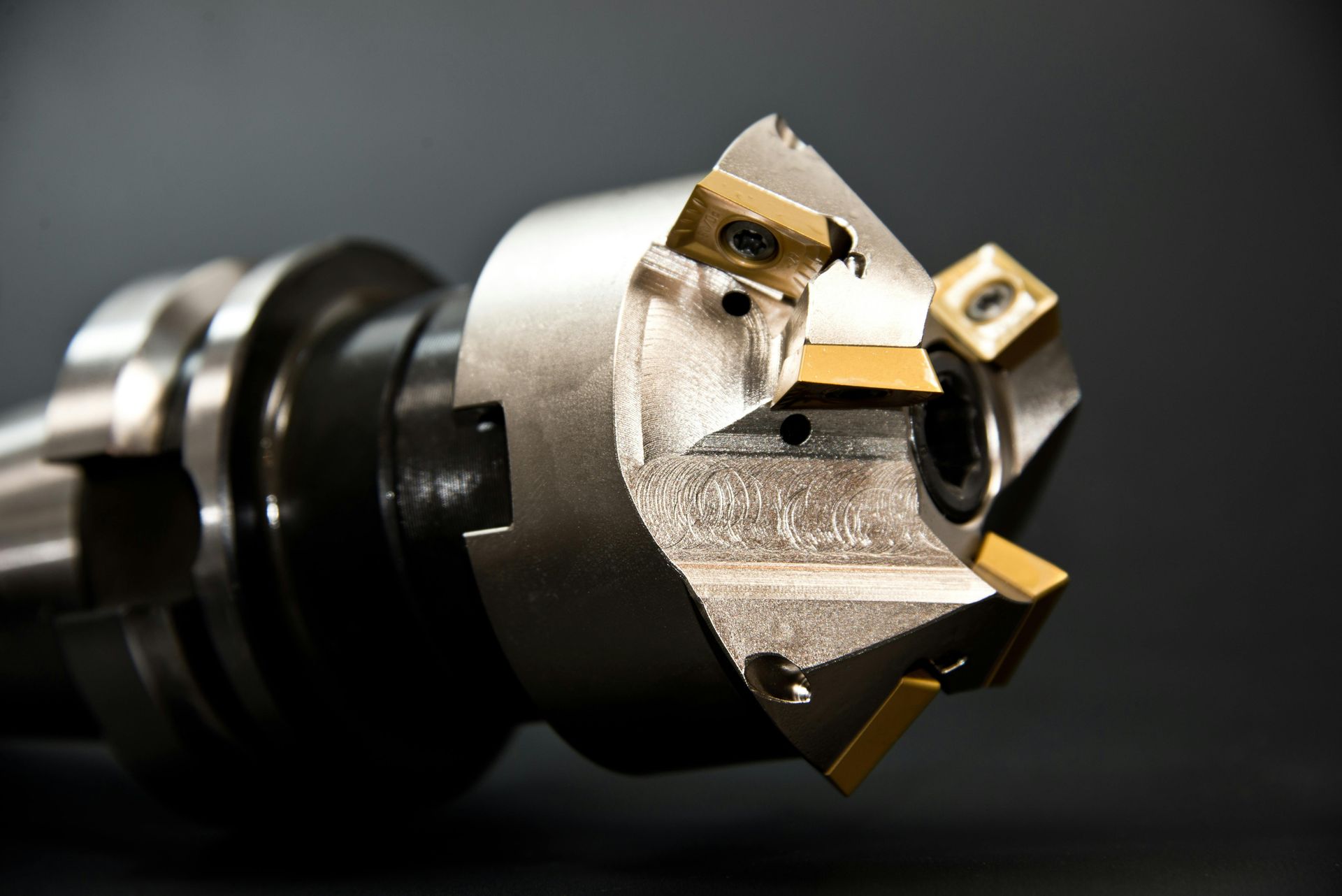 Close-up of a metal milling cutter with multiple gold-colored cutting inserts against a dark background.