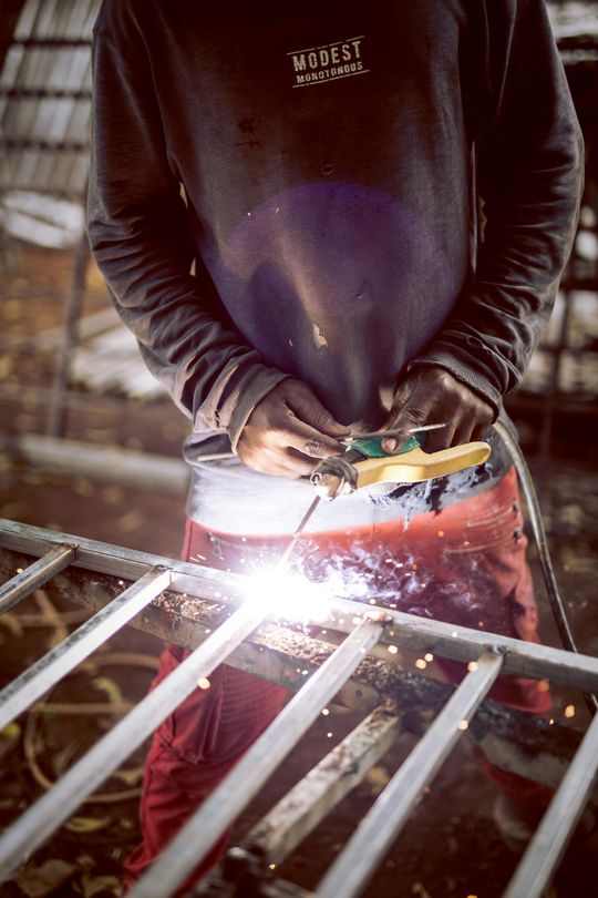 A welder in a dark shirt and red pants welding metal, sparks flying, outdoors.