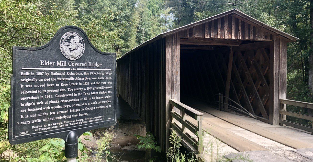 Covered bridge with a historical marker in front; wooden structure, lush forest setting.
