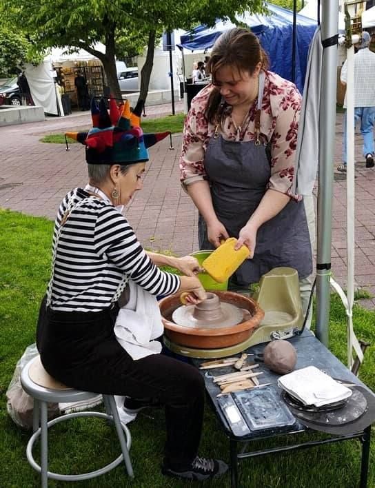 Woman at pottery wheel; another assists with a sponge, outdoors.
