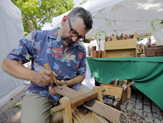 Man carving wood with a tool at an outdoor craft fair, wearing a floral shirt and glasses.