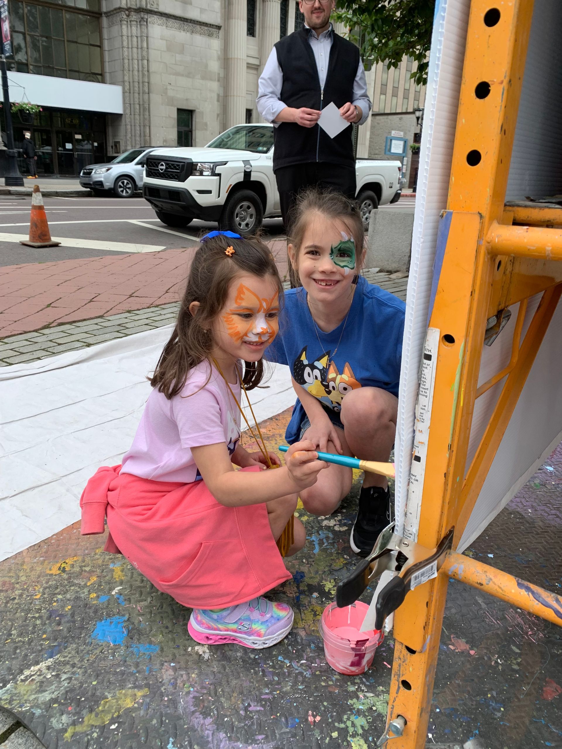 Two girls with face paint painting on a large easel outdoors. A man in the background watches.