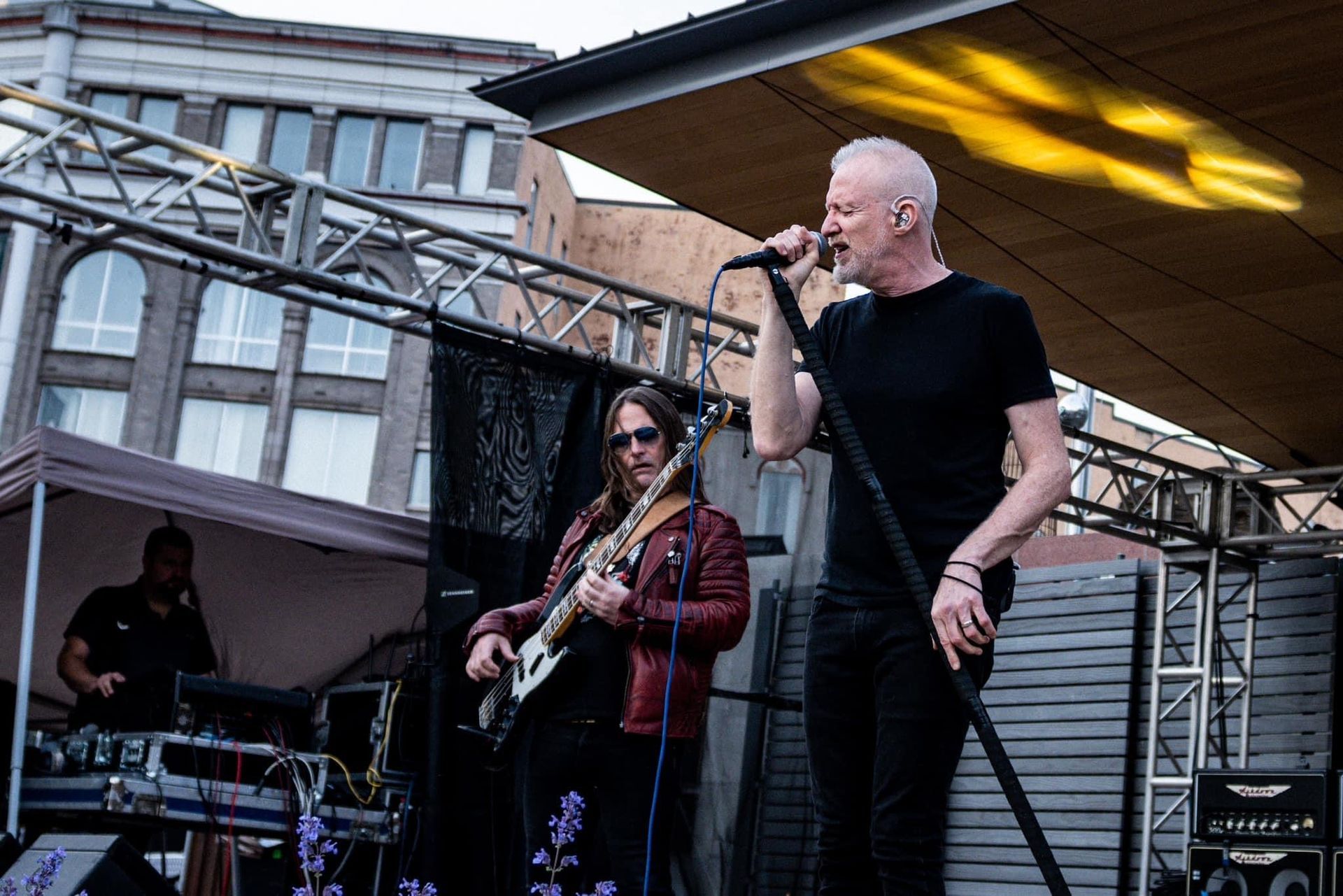 Singer on stage with band, performing outdoors. Man singing into mic, guitarist, drummer visible.
