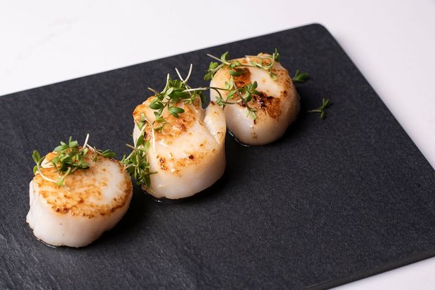 Steiktar hörpuskeljar á svörtum diski, skreyttar með grænum greinum.