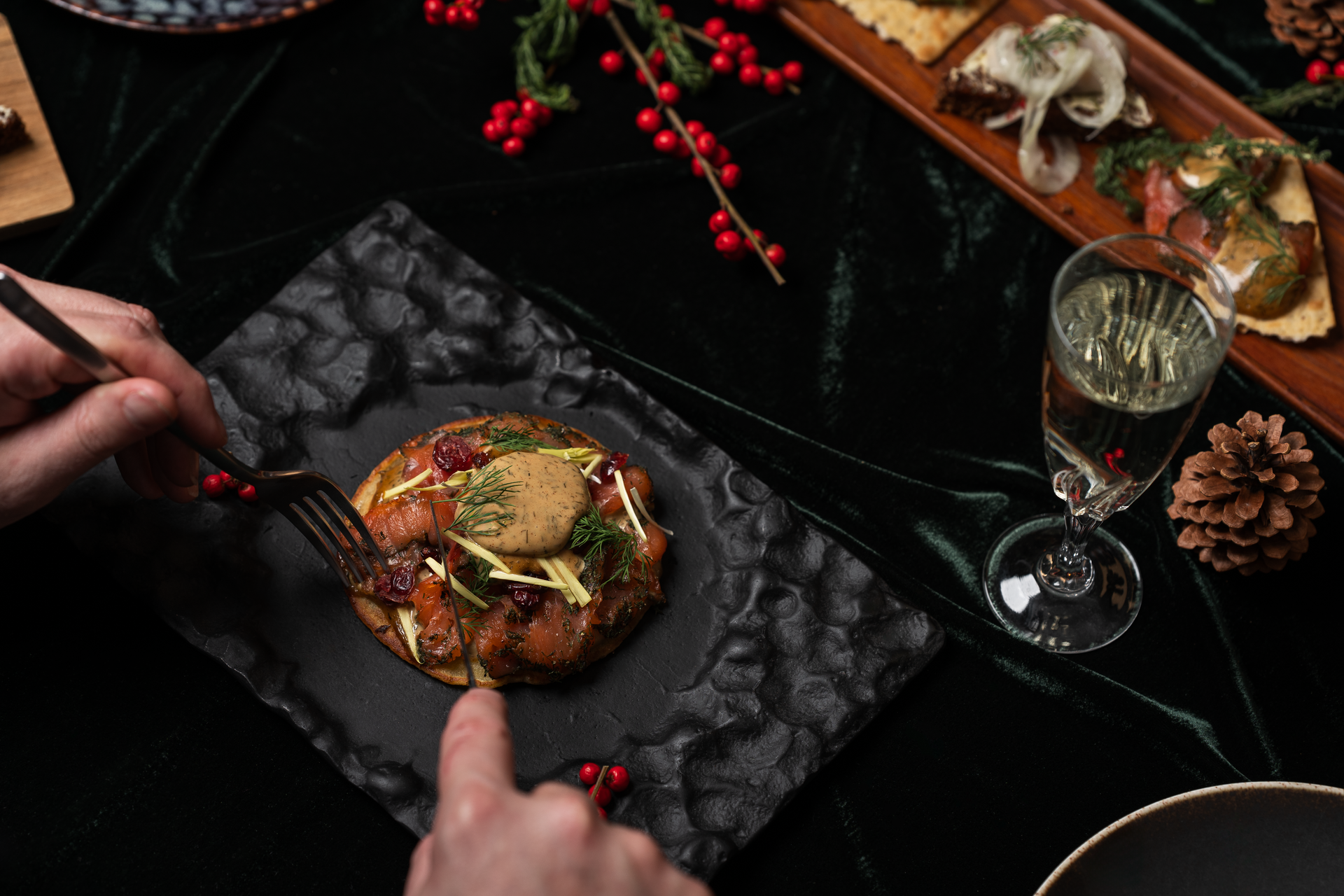 Maður notar gaffal og hníf á skreyttum diski á dökkum fleti, með glasi og öðrum forréttum í nágrenninu.