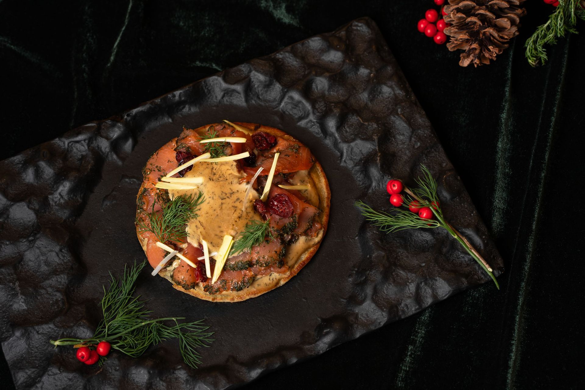 Hringlaga matur með áleggi á dökkum áferðardiski, skreyttur með berjum og furu.