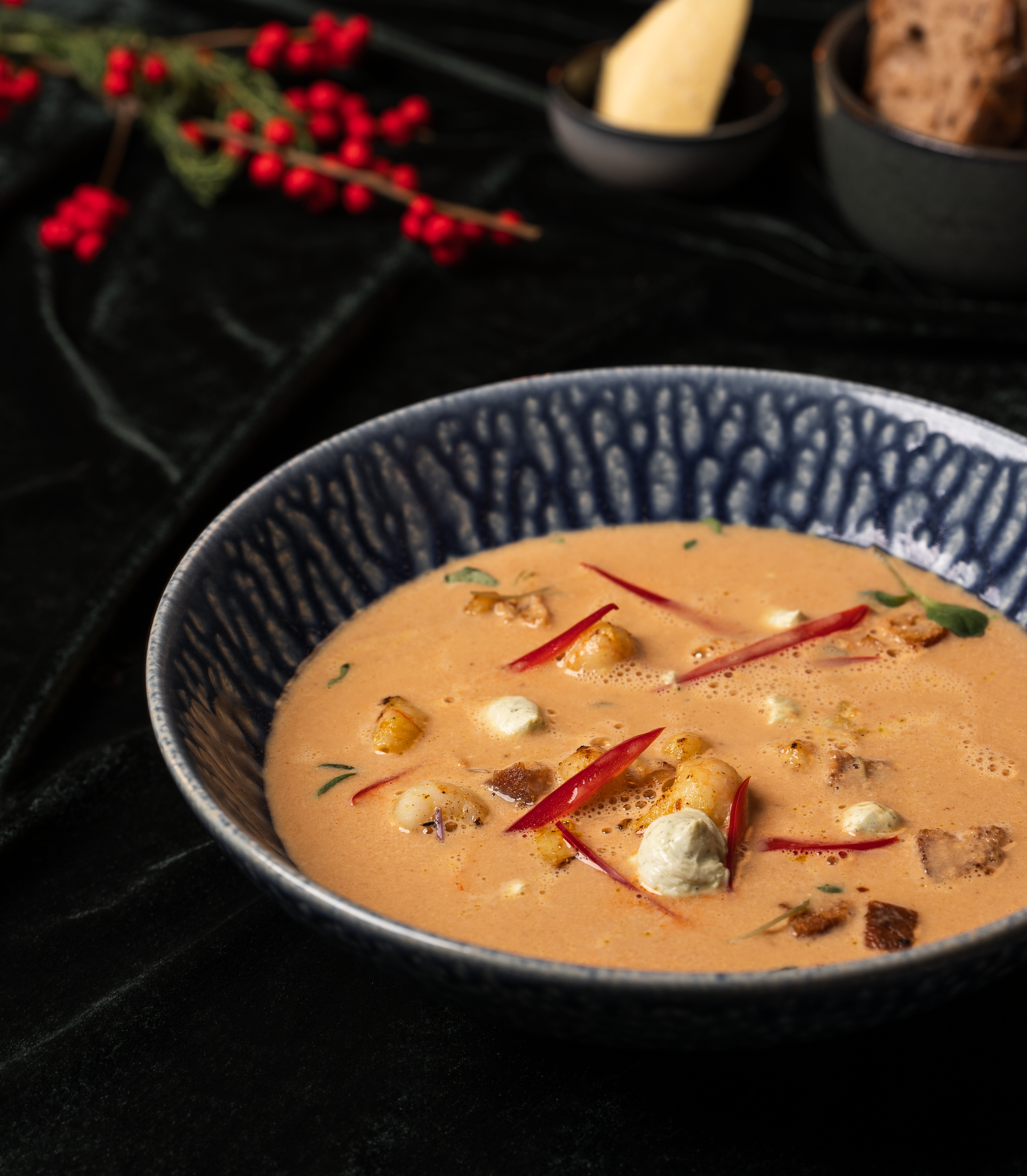 Rjómalöguð súpa í blárri skál, skreytt með rauðum paprikum og hvítum osti, á dökkum dúk.