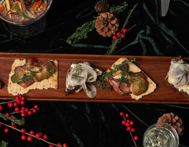 Trébakki með litlum opnum forréttum, þar á meðal súrum gúrkum, lauk og kryddjurtum, raðað á dökkgrænan flöt.