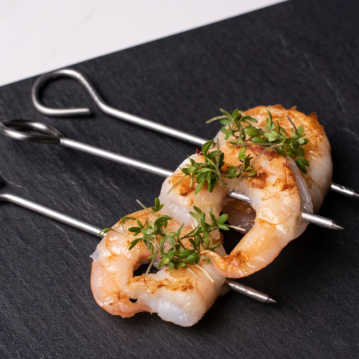 Grillaðar rækjuspjót á svörtum leirsteini, skreyttar með kryddjurtum.