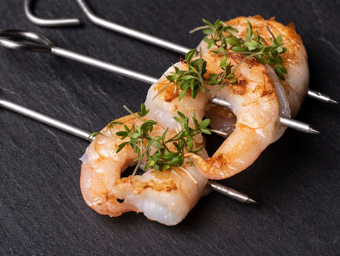 Grillaðar rækjuspjót, skreyttar með kryddjurtum, á dökkum leirflöt.