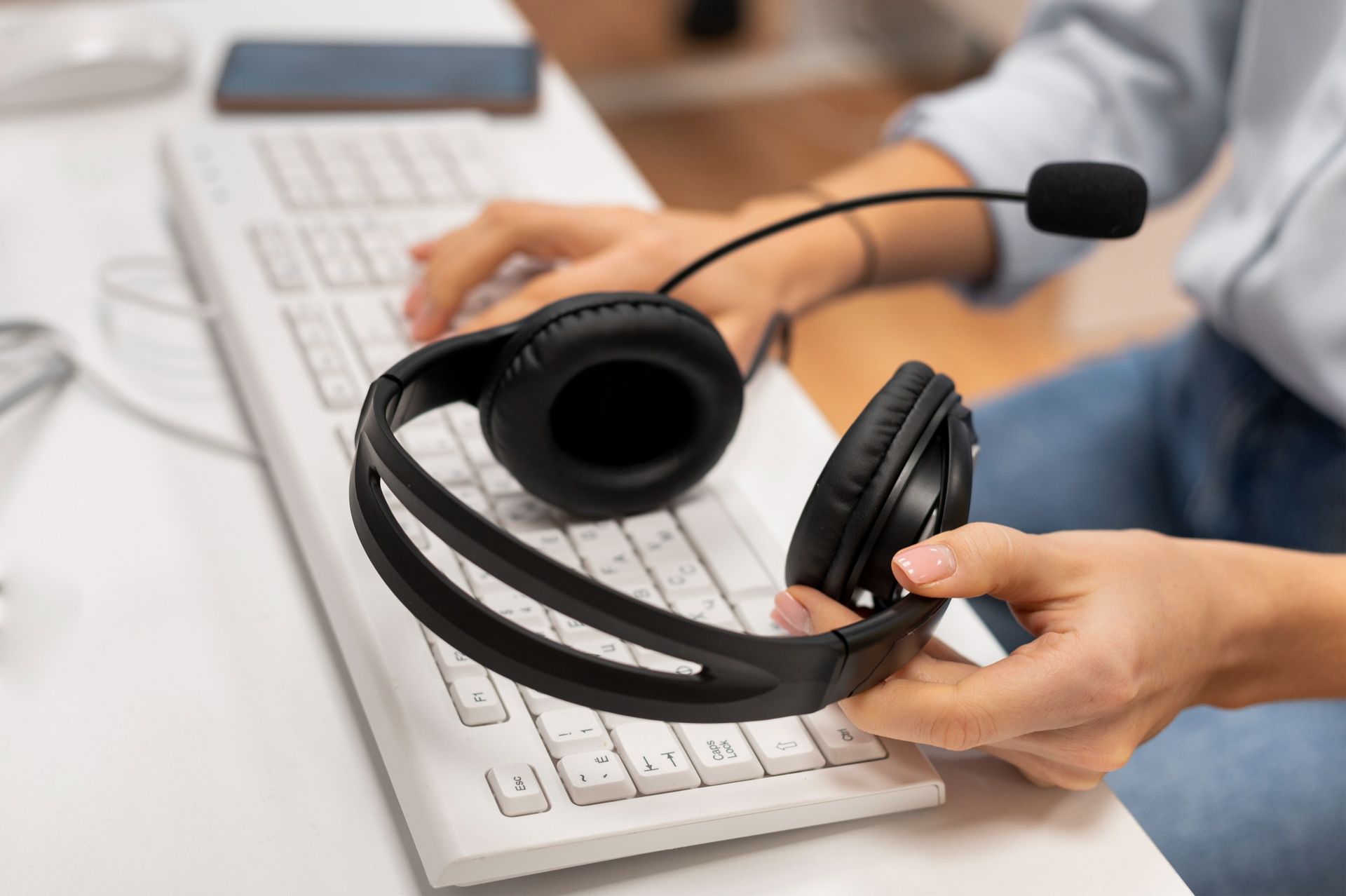 Persona escribiendo en un teclado con auriculares. El micrófono está levantado. Teclado blanco, escritorio blanco.