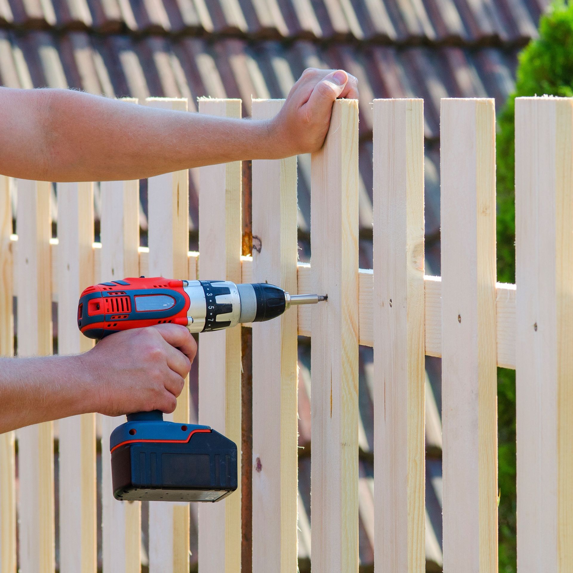 Hands using a red drill to install a white wooden fence panel outdoors