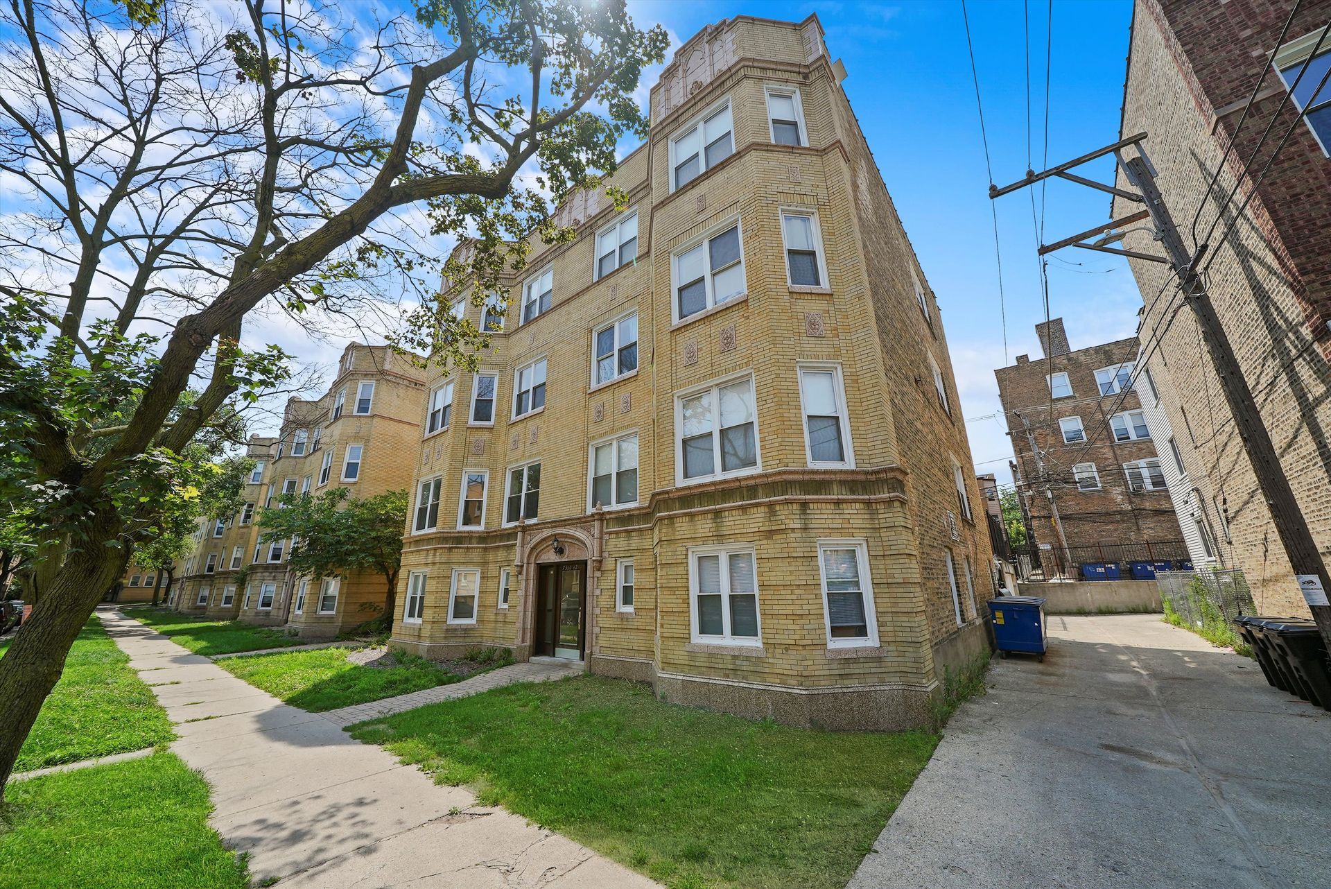 Multi-story beige brick apartment building with green lawn and tree.