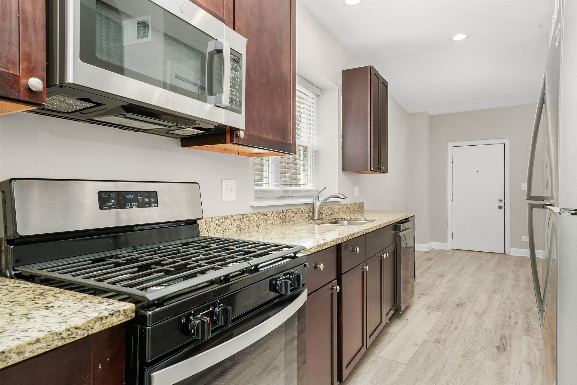 A modern kitchen with stainless steel appliances, brown cabinets, granite countertops, and light wood floors.