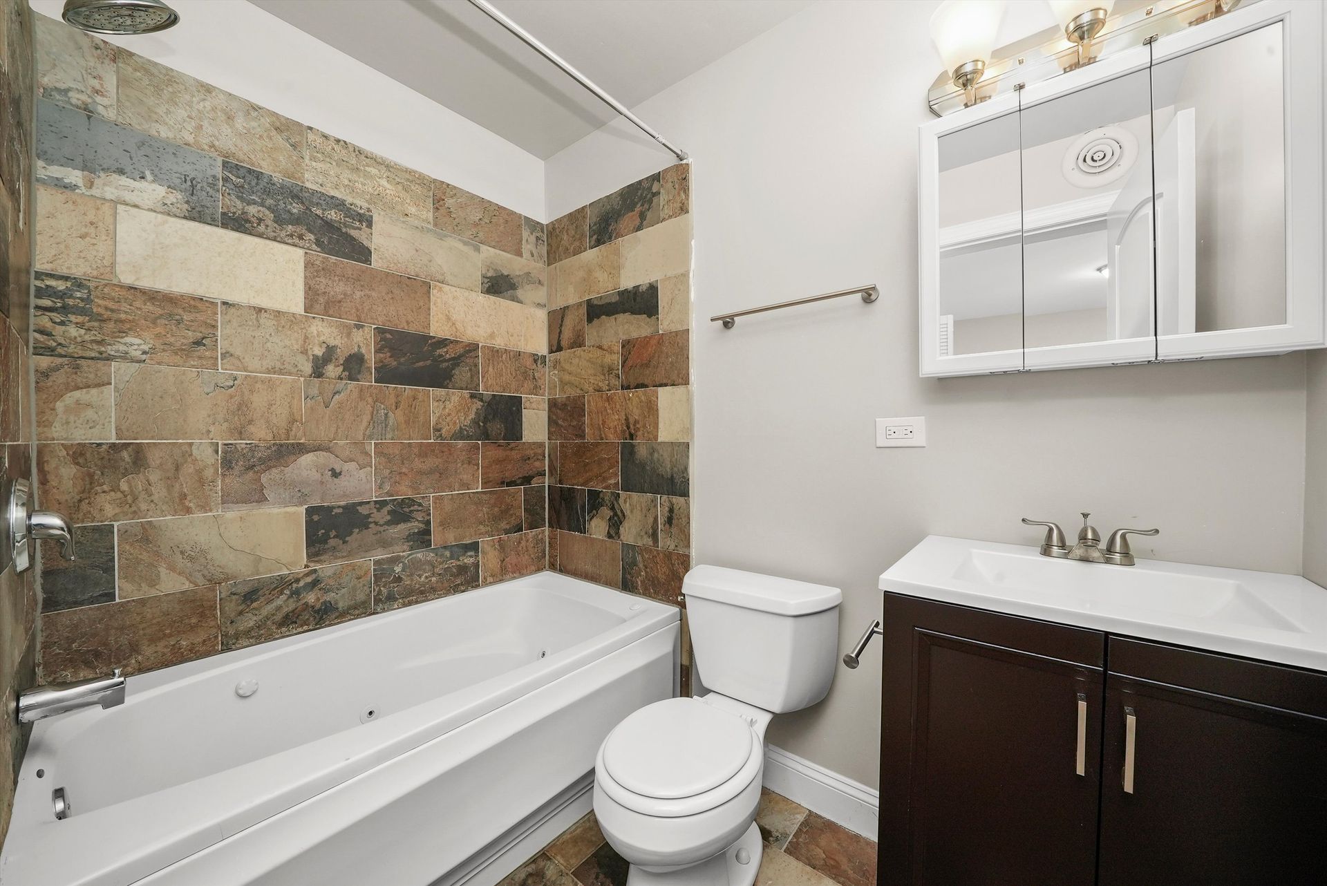 Bathroom with stone-tiled shower wall, white tub, dark vanity, toilet, and a matching floor.