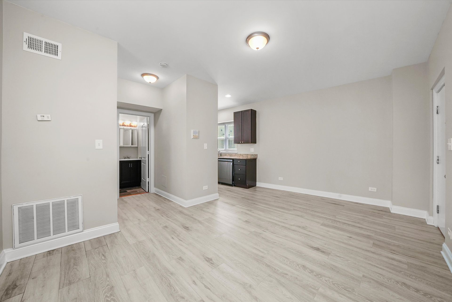 Empty, gray-walled room with wood-look flooring, doorway to a kitchen, and a small bathroom.