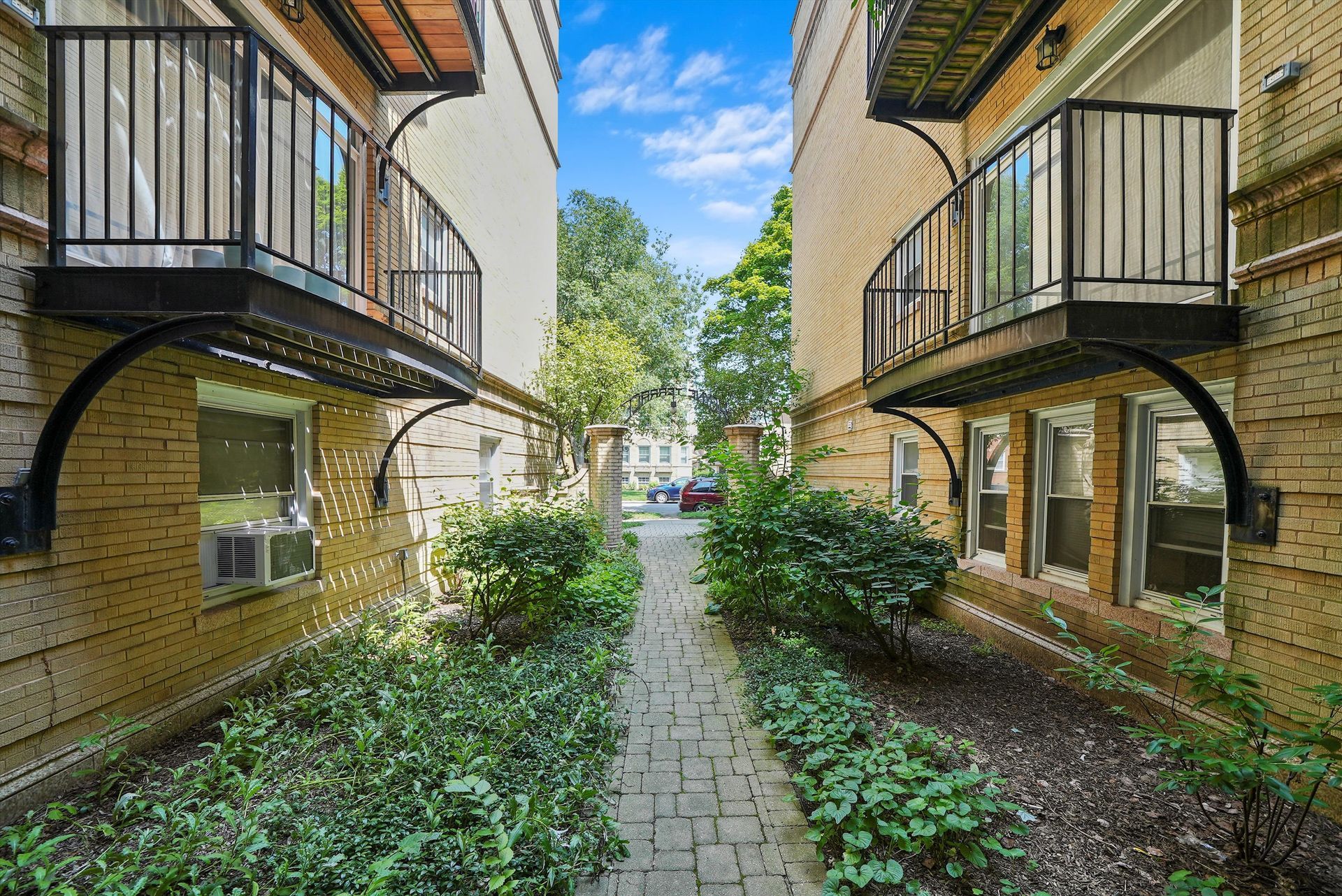 Two brick buildings with balconies flank a narrow stone path lined with greenery.