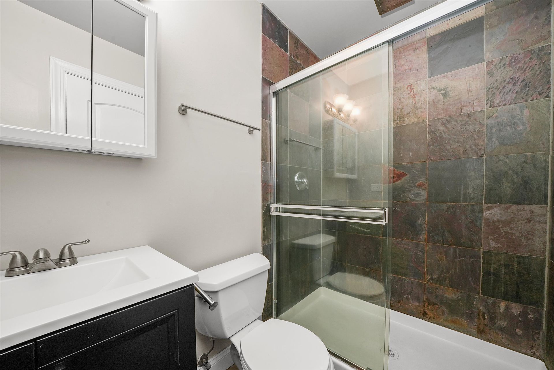 Bathroom with black vanity, white toilet, glass shower, and slate-like wall tiles.