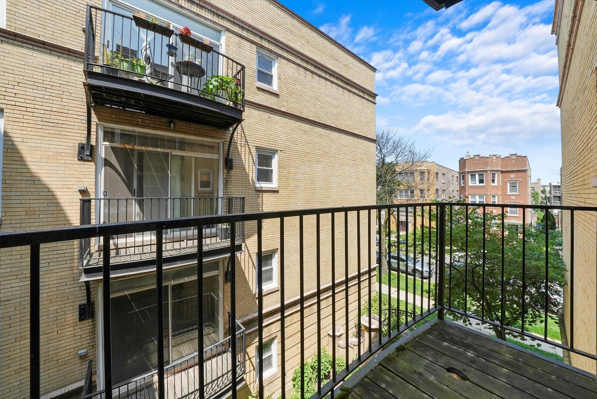 Balcony view of brick buildings, trees, and blue sky. Black metal railing, worn wood deck.