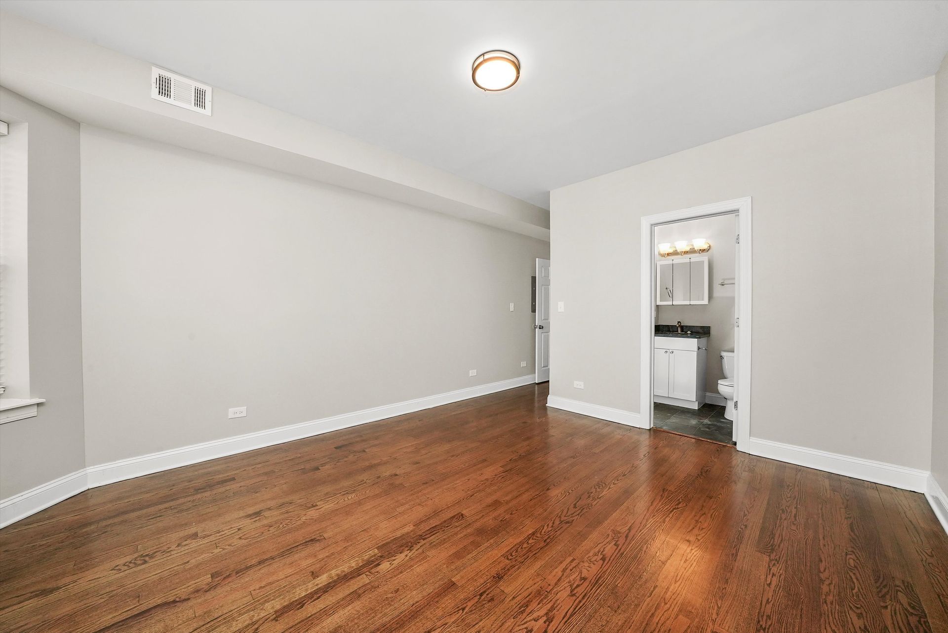 Empty room with hardwood floors, pale walls, and a bathroom doorway.