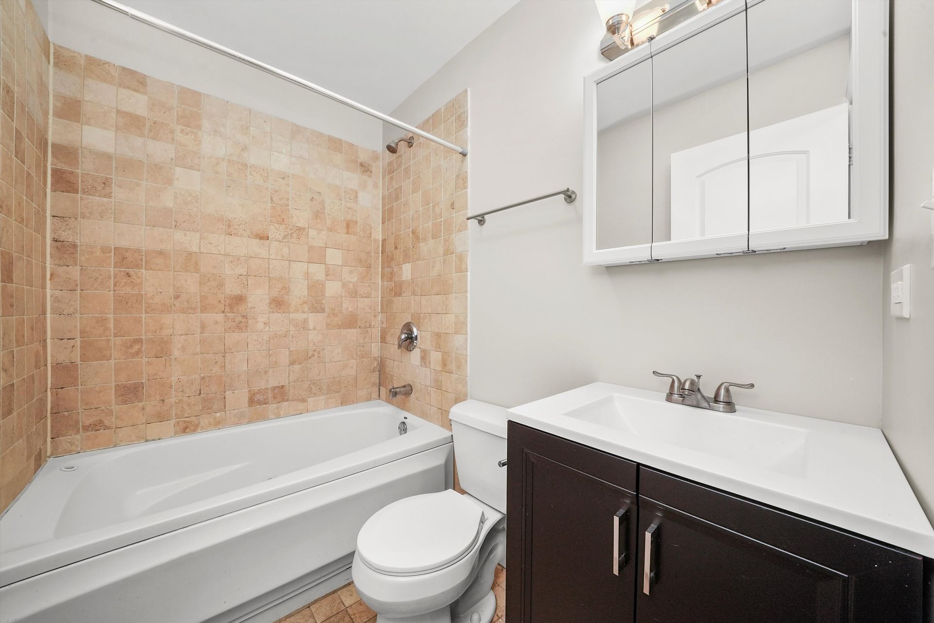 A bathroom with a bathtub, toilet, vanity with sink, and mirrored medicine cabinet. Beige tile on the walls.