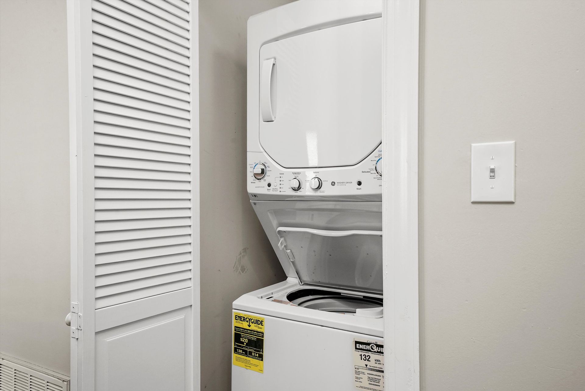 White stacked washer and dryer in a closet with a louvered door, next to a light switch.