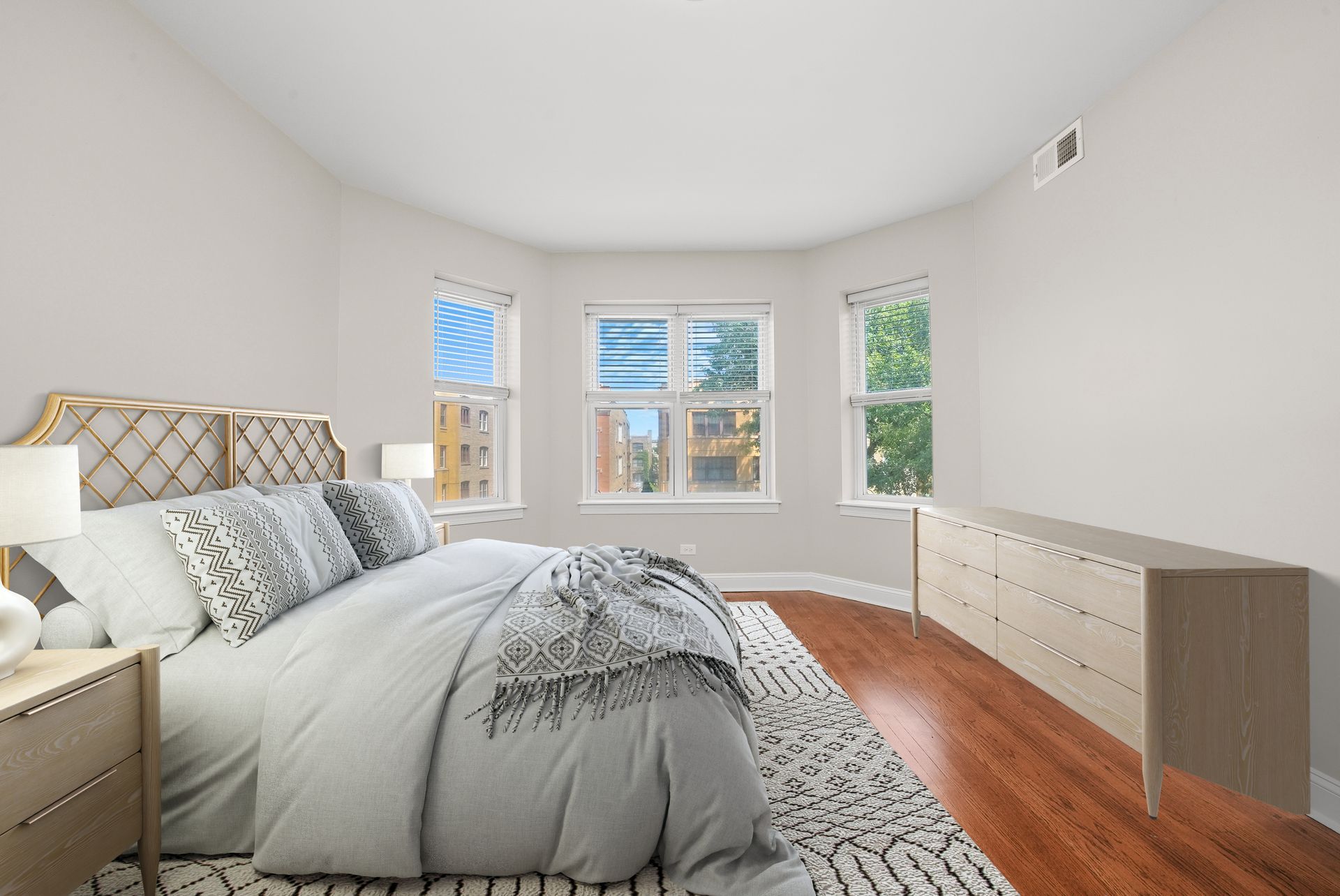 Bedroom with a bed, dresser, rug, and windows, with light-colored walls and wooden floors.