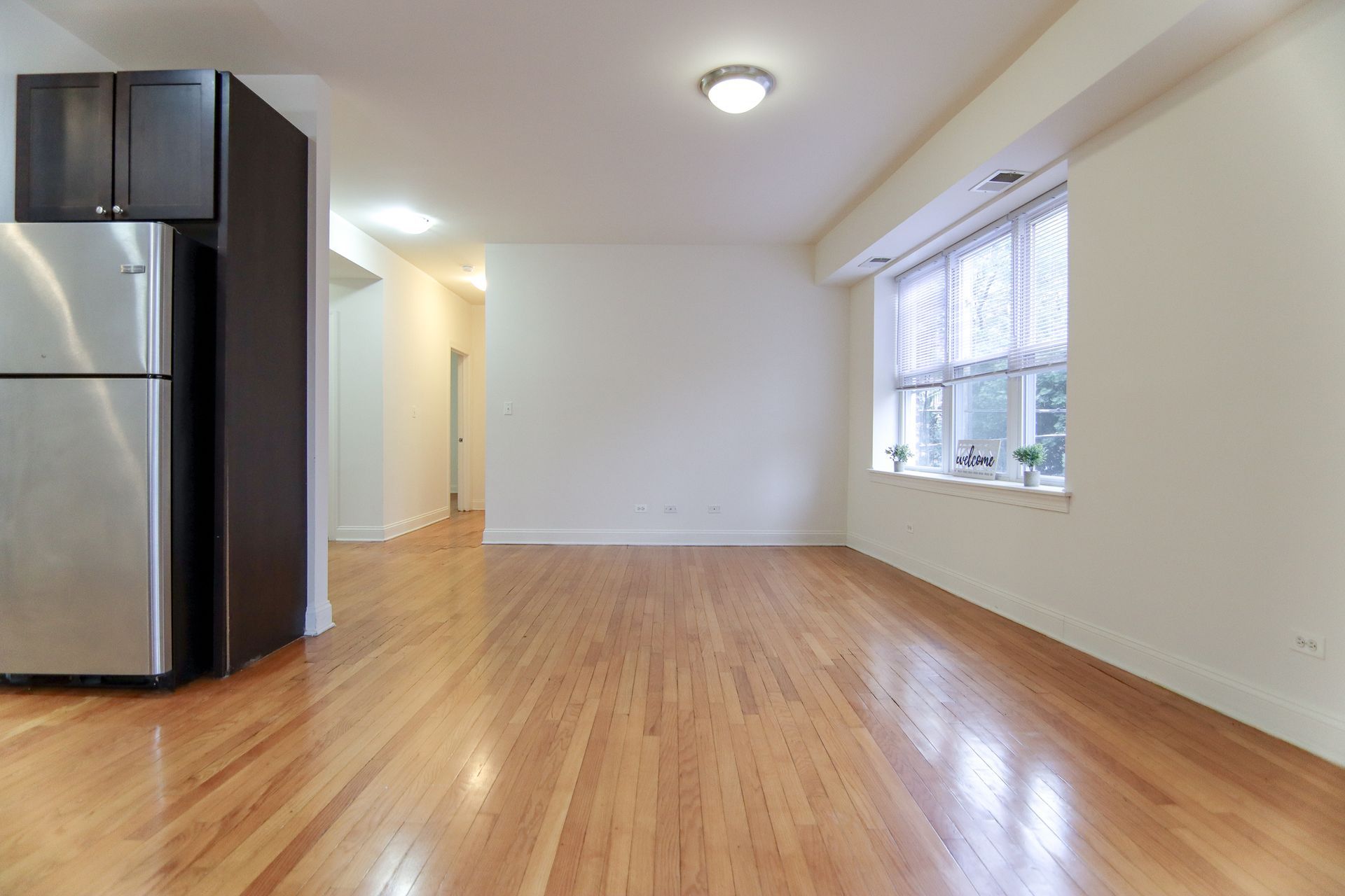 Empty room with hardwood floors, stainless steel refrigerator, and window with blinds.