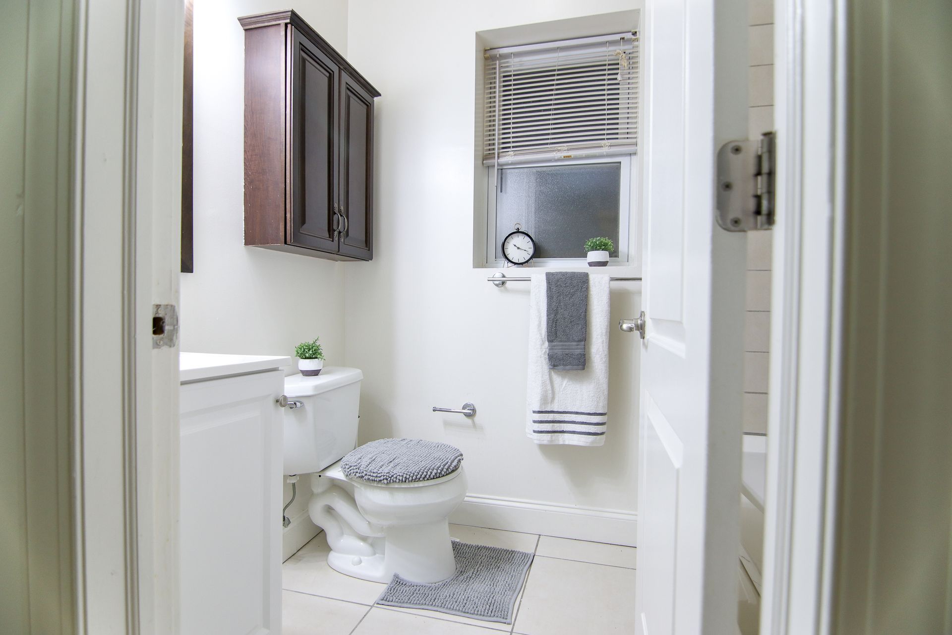 White bathroom with toilet, cabinet, window, towel, and clock.