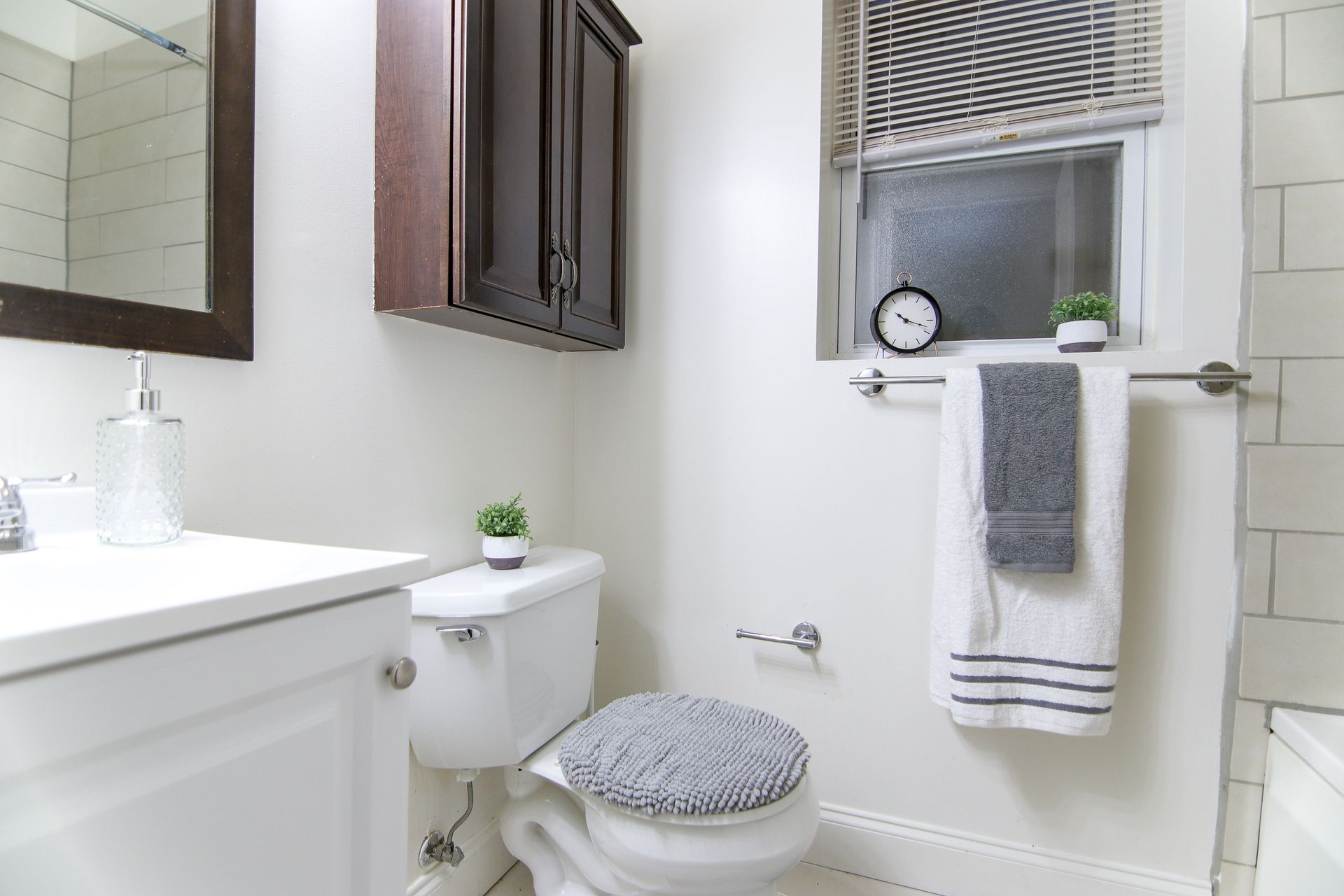 Bathroom with white sink, toilet, and walls; dark brown cabinet; gray towels; small clock and plants.